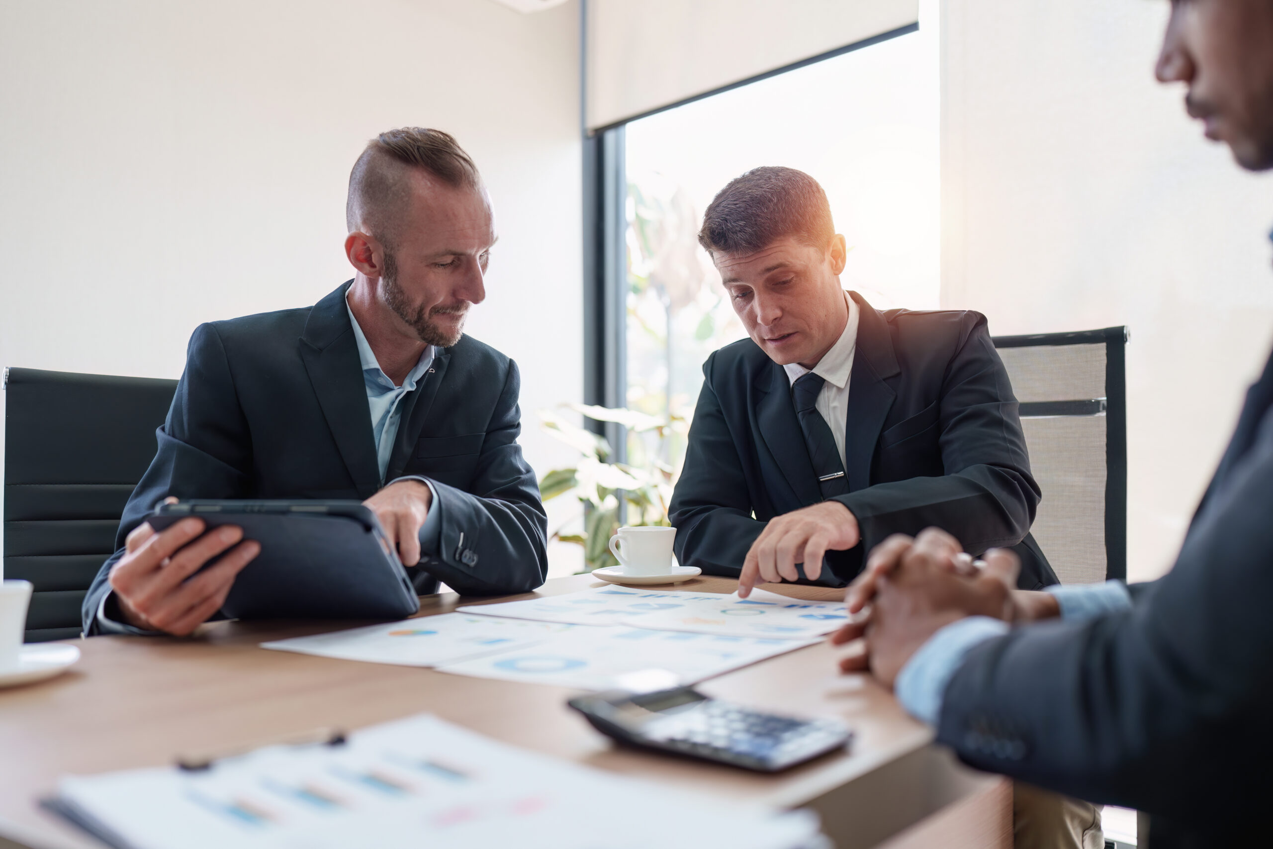 Business professionals in formal attire discussing charts and graphs of offshore trust setup at a meeting table, with a tablet and documents spread out in a bright office setting.